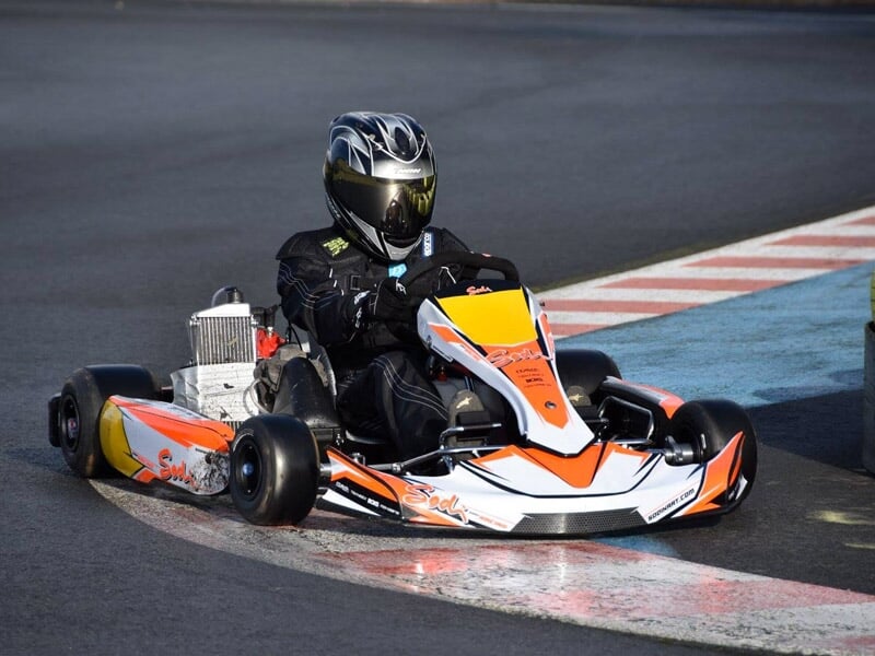 Pilote en combinaison noire et casque intégral conduisant un karting orange et blanc sur piste asphaltée en virage.