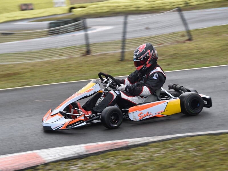Conducteur en tenue de course et casque rouge au volant d'un karting orange et blanc sur une piste avec herbe en arrière-plan.