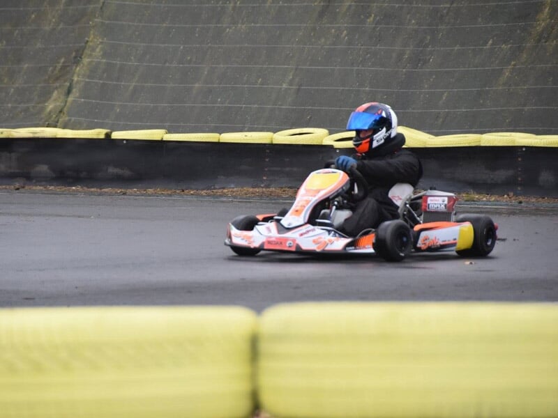 Pilote avec casque bleu et combinaison noire en train de piloter un karting orange et blanc sur une piste de karting.