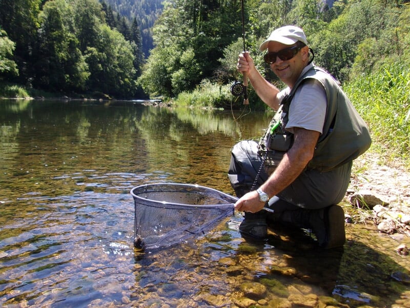 Un pêcheur souriant en tenue de pêche à la mouche tient une épuisette dans une rivière ensoleillée.