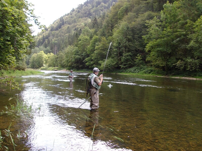 Deux pêcheurs en waders pêchent à la mouche dans une rivière entourée de forêts verdoyantes.