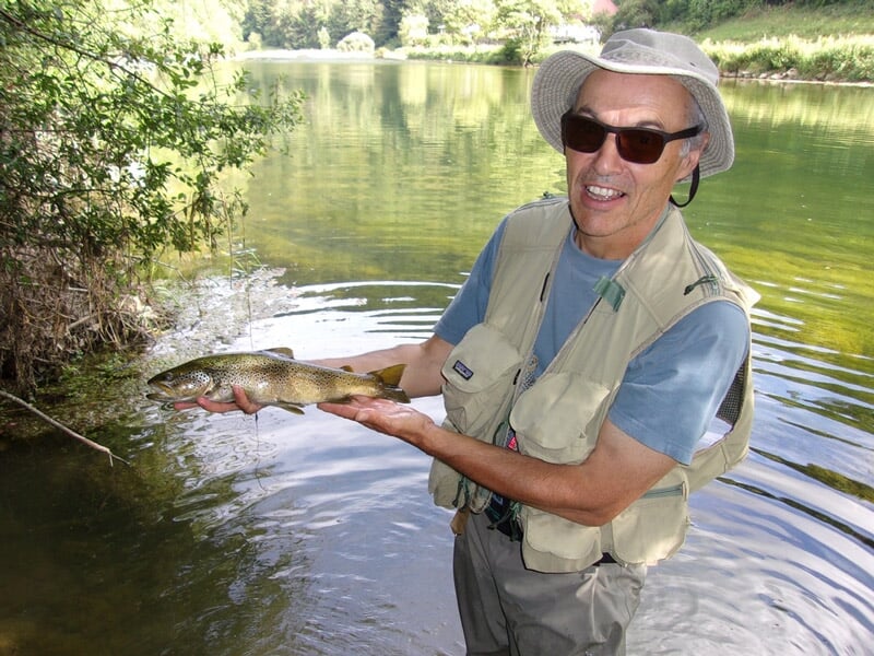 Un pêcheur souriant en chapeau et lunettes de soleil tient une truite dans une rivière calme entourée de verdure.