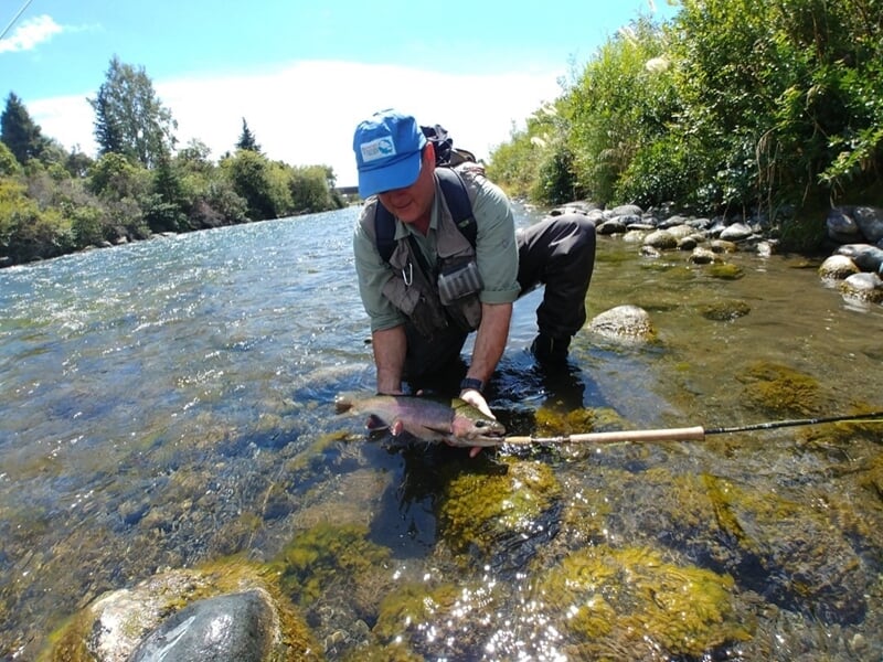 Pêcheur agenouillé dans une rivière, relâchant délicatement un poisson dans l'eau sous un ciel bleu.