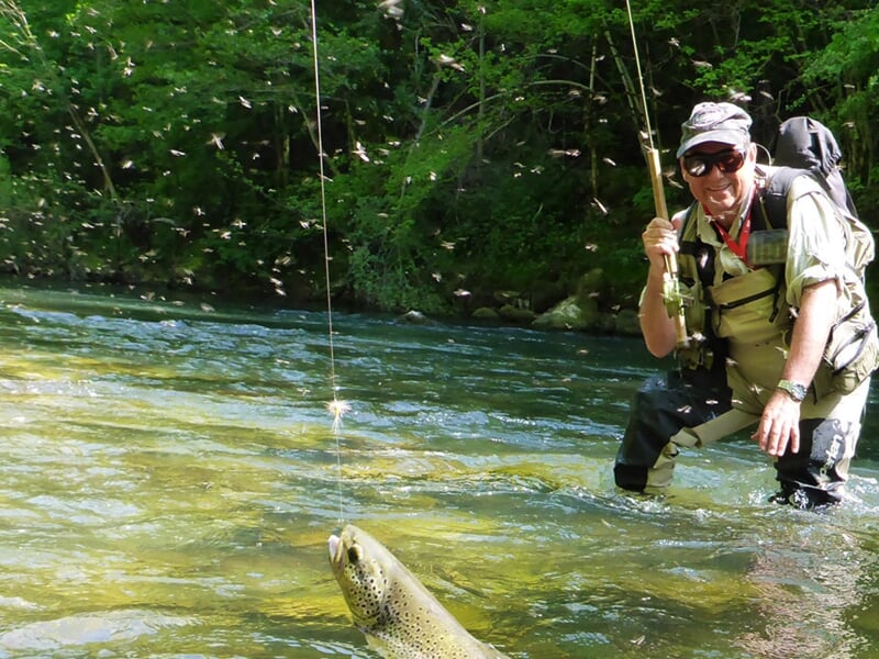 Pêcheur souriant dans une rivière, tenant une canne à pêche avec un poisson suspendu au bout de la ligne.