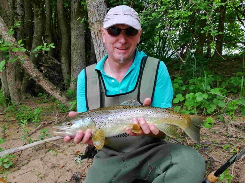 Homme souriant en tenue de pêche tenant une truite brune fraîchement pêchée au bord d'une rivière boisée.