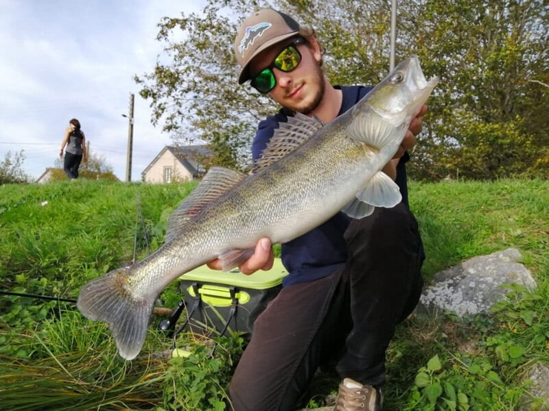 Pêcheur en lunettes de soleil tenant un grand poisson argenté avec un paysage vert en arrière-plan.