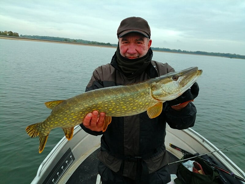 Homme souriant en tenue chaude tenant un grand brochet sur un bateau au milieu d'un lac calme.