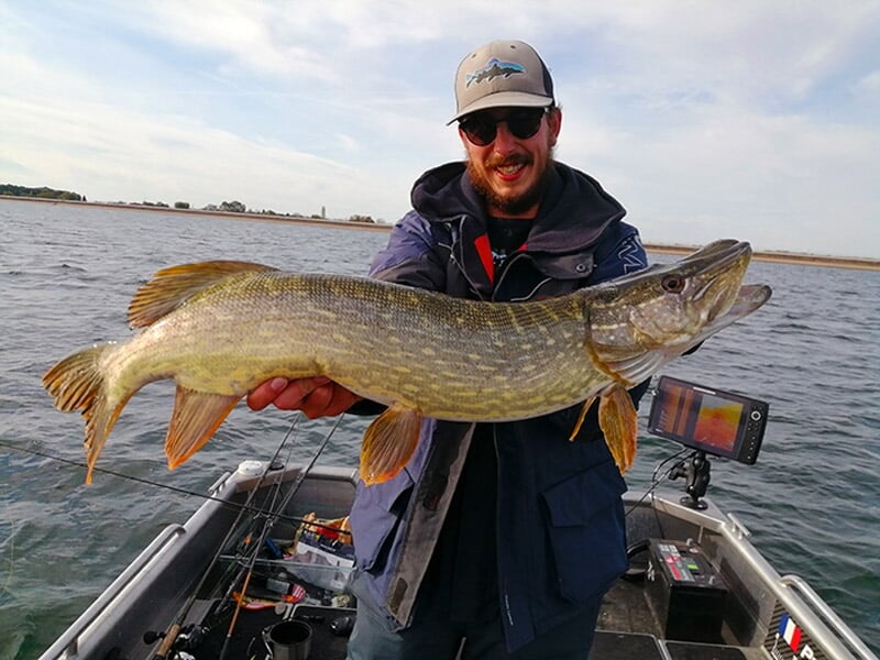 Pêcheur en veste bleue tenant un brochet impressionnant sur un bateau équipé de matériel de pêche.