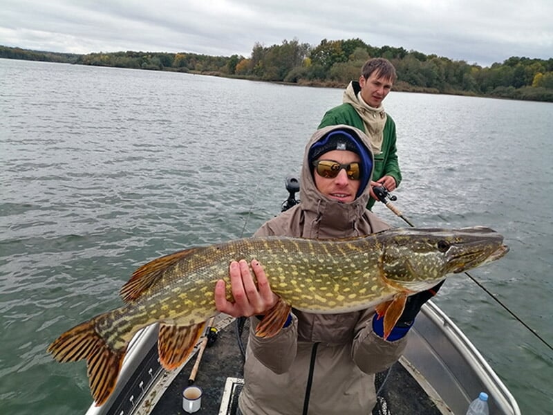 Deux pêcheurs sur un bateau tenant un brochet, souriant sous un ciel nuageux au-dessus d'un lac.