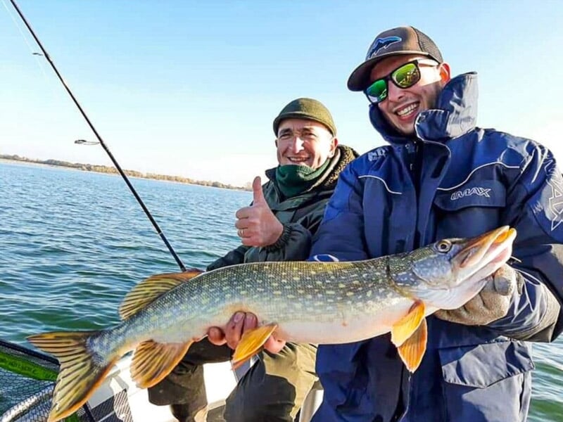Deux hommes en vêtements chauds sur un bateau tenant un brochet, l'un fait un pouce levé en signe de succès.