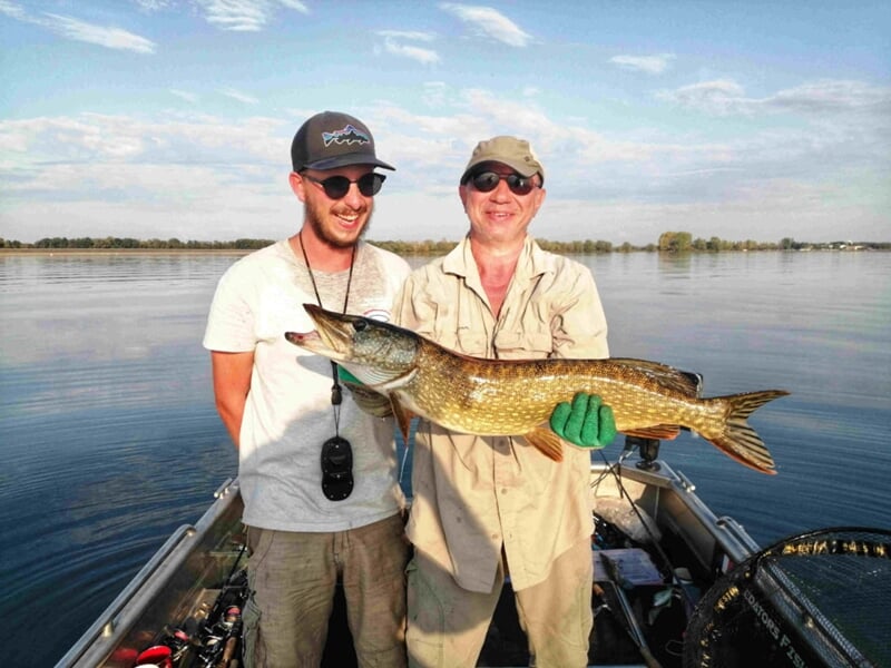 Deux pêcheurs souriants sur un bateau tenant un brochet, avec un lac calme et un ciel clair en arrière-plan.