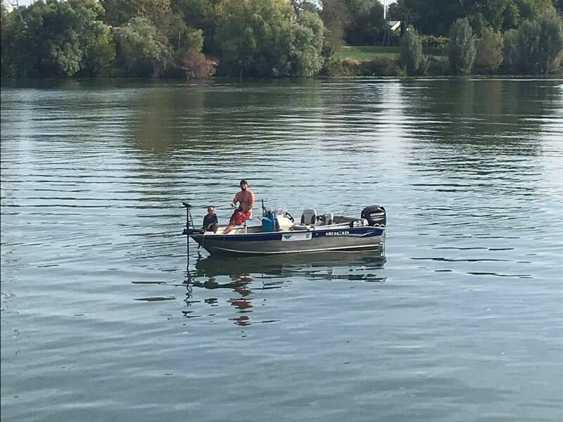Deux pêcheurs sur un bateau au milieu d'une rivière calme entourée d'arbres verts en arrière-plan.