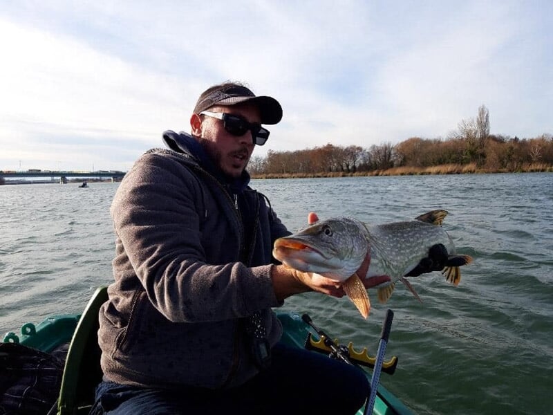 Un homme en veste et casquette tenant un gros poisson fraîchement pêché sur un bateau en rivière.
