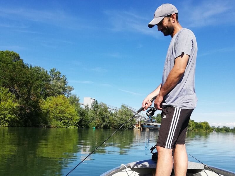 Un homme en tenue décontractée pêche au lancer depuis un bateau sur une rivière bordée d'arbres.