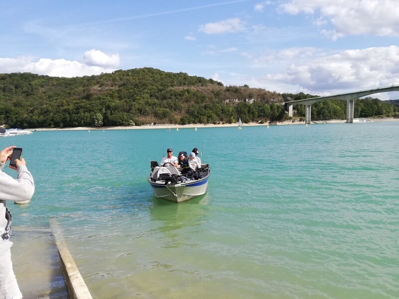 Un bateau avec plusieurs personnes navigue sur une eau turquoise près d'un pont et d'une rive boisée.