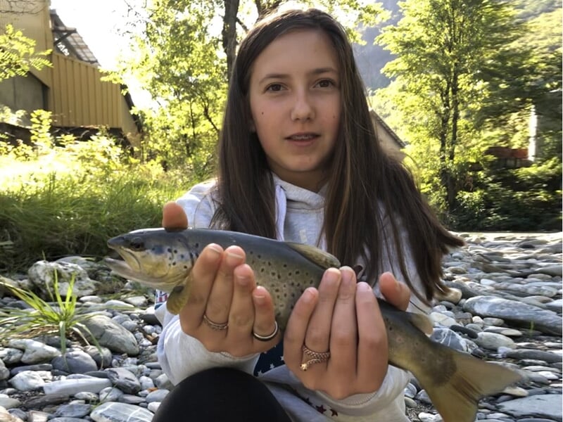 Jeune fille assise au bord d'une rivière, tenant une truite avec des rochers et de la verdure en arrière-plan.
