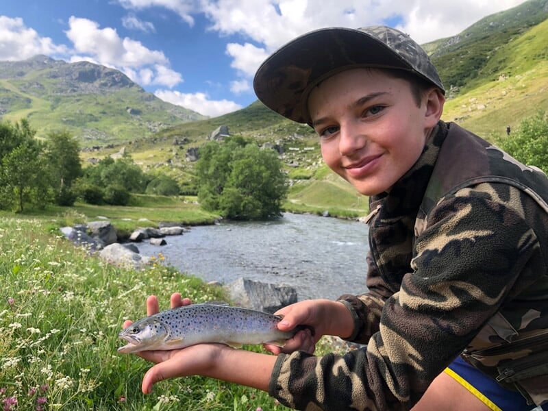 Garçon souriant tenant une truite au bord d'une rivière, paysage de montagne et ciel bleu en arrière-plan.