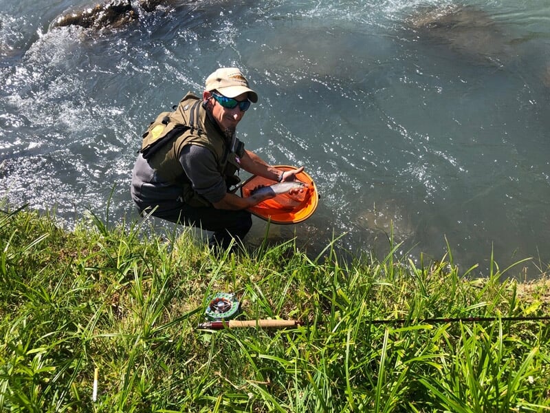 Pêcheur accroupi au bord d'une rivière, tenant une épuisette orange dans l'eau, équipement de pêche visible.