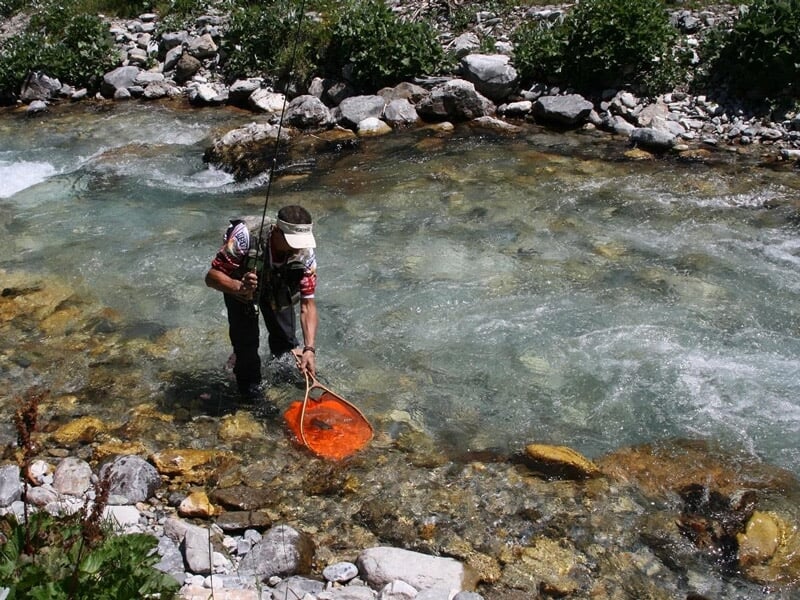 Pêcheur dans une rivière de montagne avec une épuisette orange, eau claire et rochers visibles.