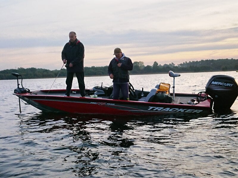 Deux pêcheurs debout sur un bateau rouge pêchent sur un lac calme avec un ciel nuageux en arrière-plan.
