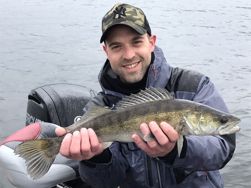 Un homme souriant montre un poisson sandre qu'il tient à deux mains, vêtu d'une veste grise et casquette camouflage.