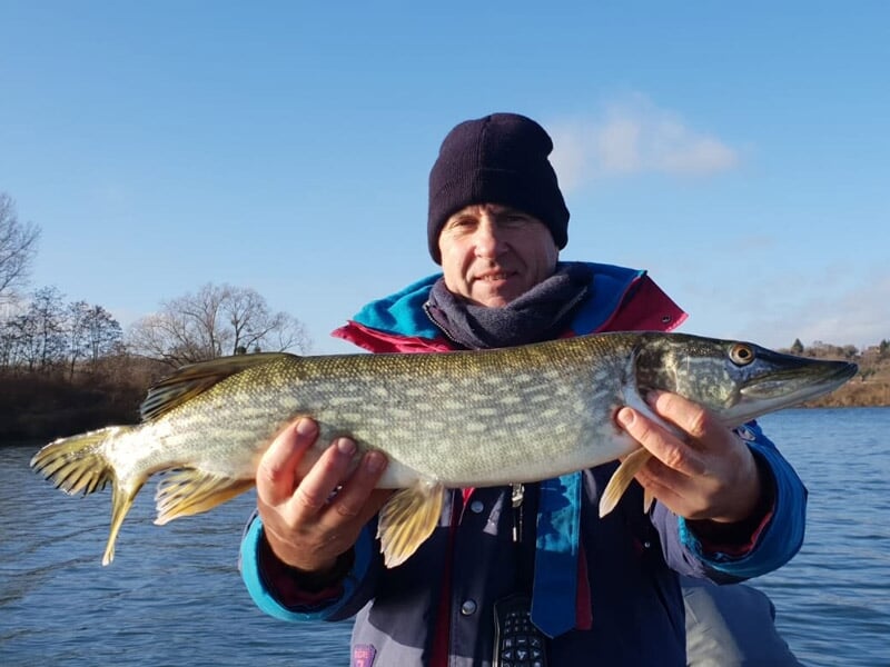 Un homme en bonnet noir et veste bleue tient un brochet de taille moyenne devant lui, au bord d'un lac.