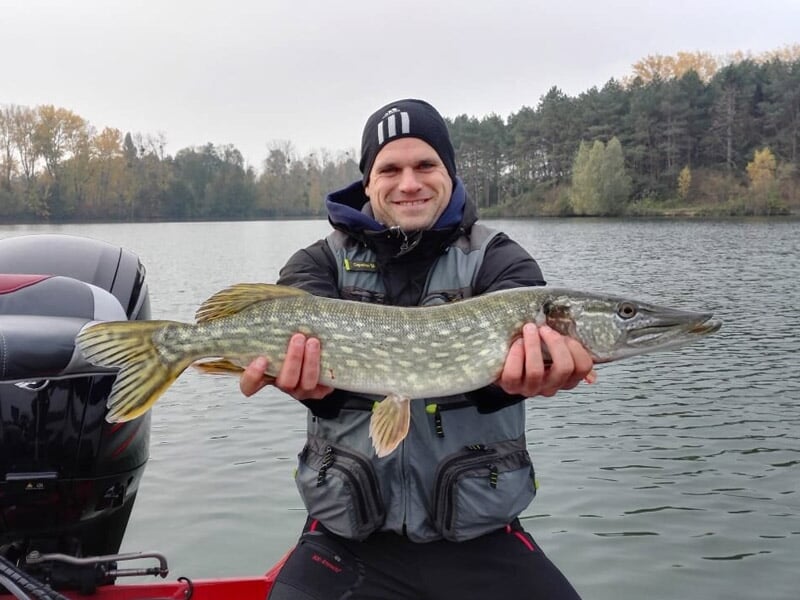 Un pêcheur souriant en gilet de sauvetage tient un brochet allongé devant un lac entouré d'arbres en automne.