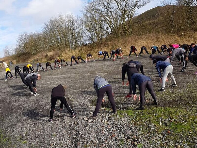 Groupe de personnes effectuant des exercices d'étirement en cercle sur un sol caillouteux.