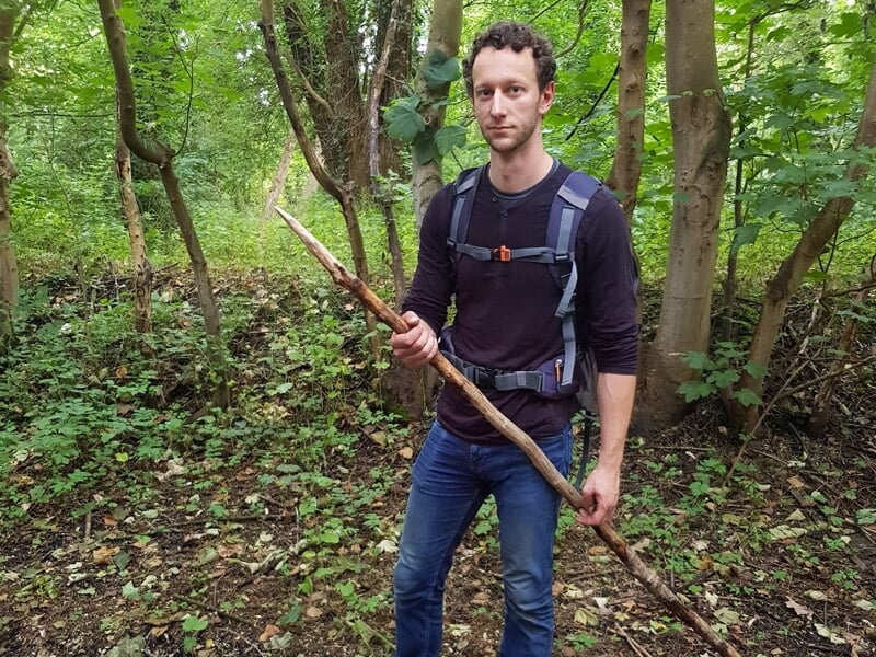 Homme tenant un bâton dans une forêt dense, prêt pour une activité de survie ou randonnée.