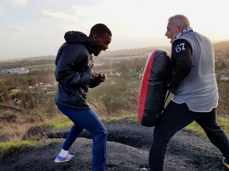 Deux hommes s'entraînant au combat avec un bouclier et des gants dans un cadre naturel.