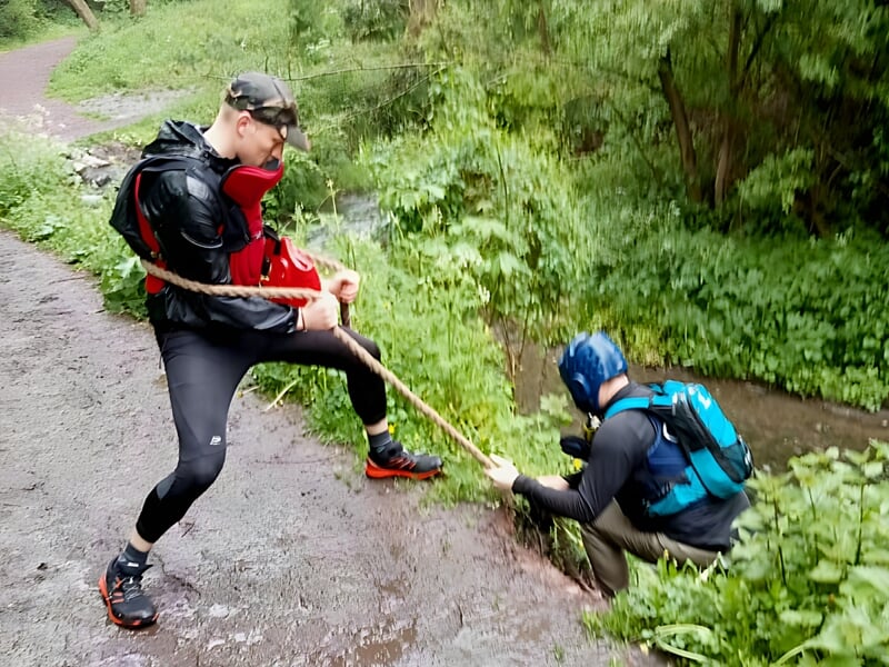 Deux hommes en tenue de sport participent à un stage commando, l'un aide l'autre à remonter d'un fossé avec une corde.