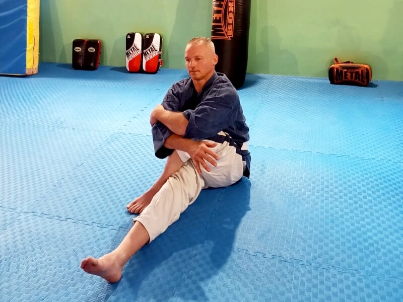 Homme assis sur un tapis bleu en tenue d'arts martiaux, effectuant un étirement.
