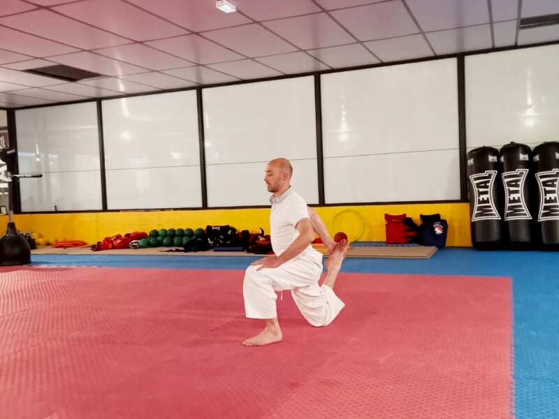 Homme en kimono blanc réalisant un exercice d'étirement sur un tapis rouge en salle.