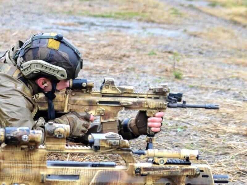 Soldat en tenue camouflage allongé au sol, visant avec un fusil d'assaut lors d'un stage commando en extérieur.