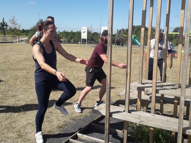 Trois personnes participent à un parcours d'obstacles en plein air, avec des structures en bois et un sol sec.