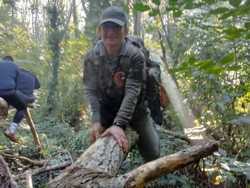 Une femme en tenue camouflage grimpe sur un tronc d'arbre dans une forêt dense et ensoleillée.
