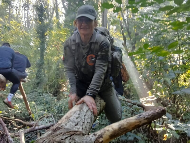 Une femme en tenue camouflage grimpe sur un tronc d'arbre dans une forêt dense et ensoleillée.