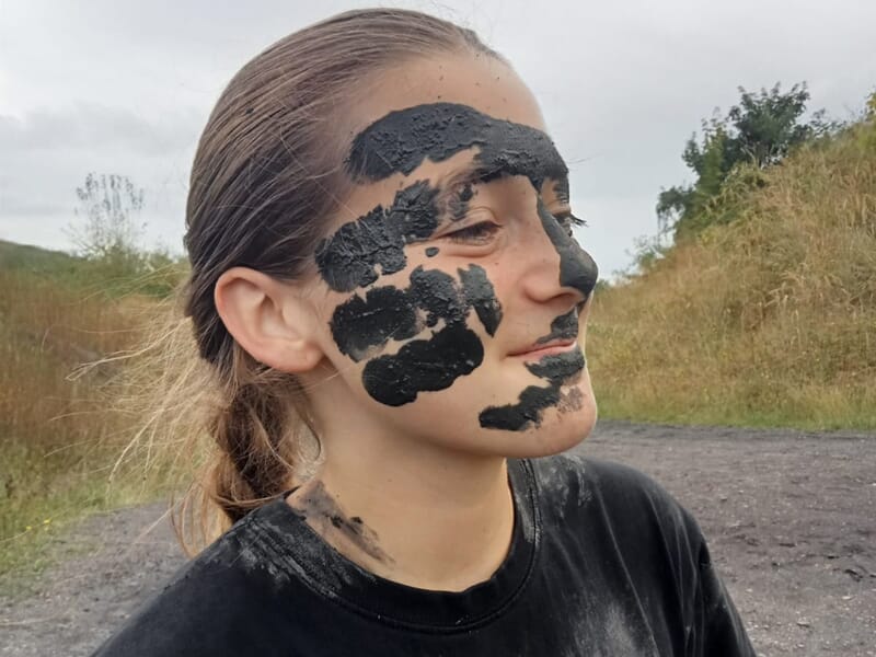 Portrait d'une jeune femme avec de la boue noire peinte sur le visage, en extérieur sur un terrain naturel.