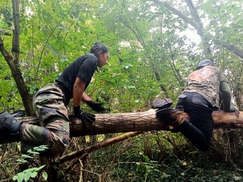 Deux personnes franchissent un obstacle en bois dans une forêt, participant à un stage commando exigeant.