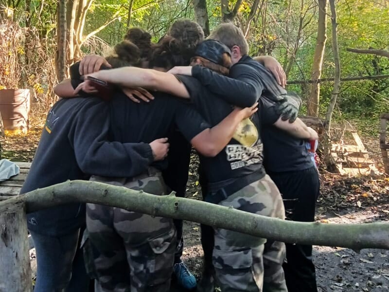 Un groupe de personnes en tenue militaire se serre dans les bras en cercle dans une forêt, montrant l'esprit d'équipe.