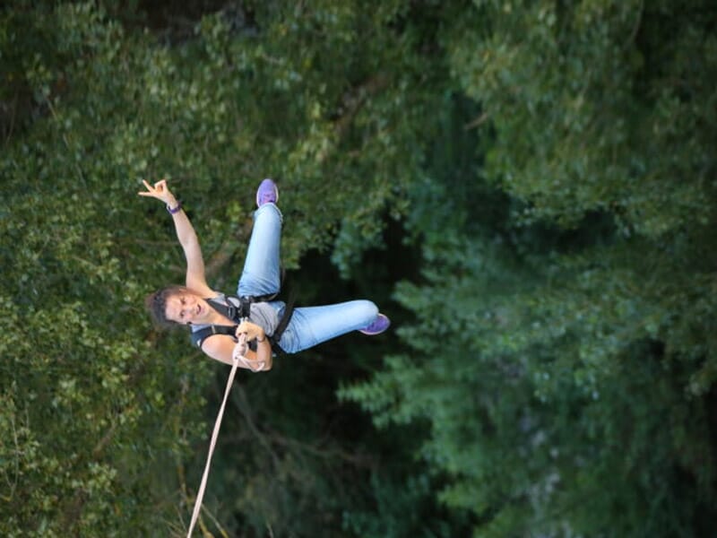 Saut à l'élastique en pleine nature avec une femme en jeans, suspendue dans les airs, souriante et faisant un signe de la main. En arrière-plan, une forêt dense et verdoyante.