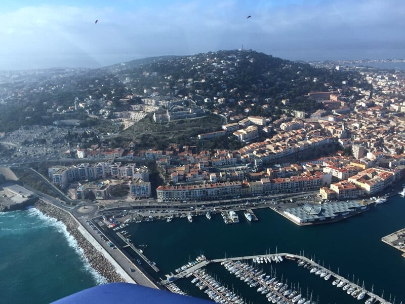 Vue aérienne d'une ville côtière avec un port de plaisance, des collines et un ciel clair.