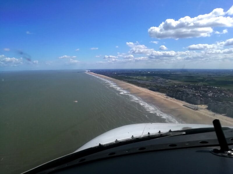 Vue depuis le cockpit d'un avion survolant une longue plage bordée par la mer et un ciel partiellement nuageux.