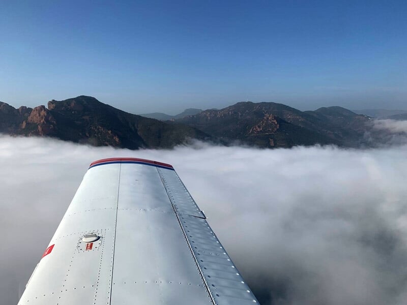 Aile d'un avion survolant une mer de nuages avec des montagnes visibles au loin sous un ciel bleu.