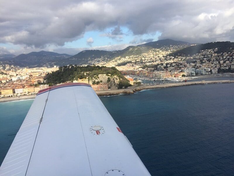 Aile d'un avion survolant une ville côtière avec des collines et une mer calme sous un ciel clair.