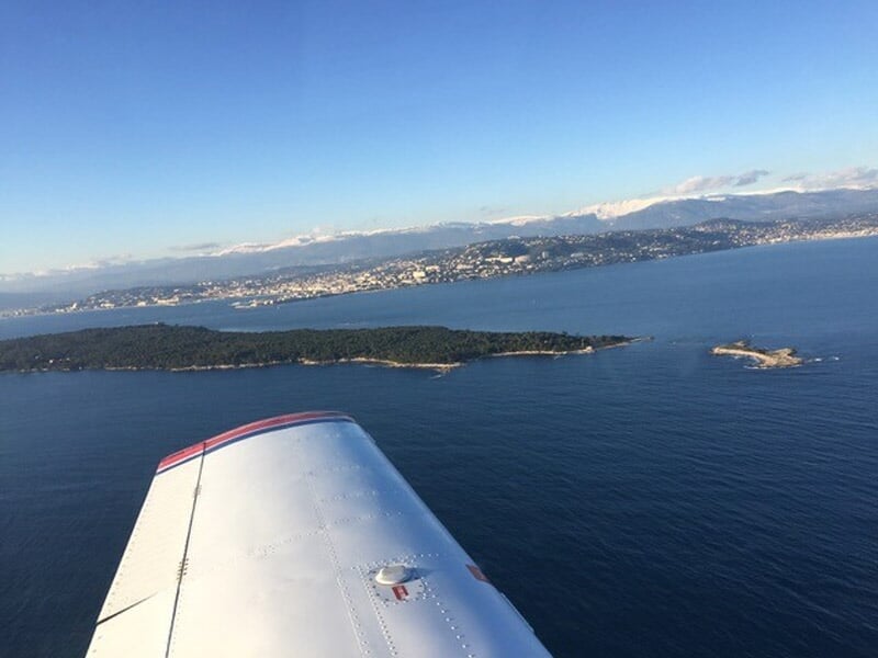 Aile d'un avion survolant une île entourée d'eau bleue avec un ciel dégagé et quelques nuages.