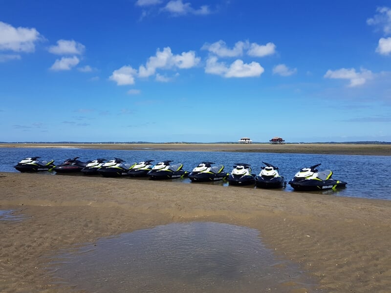 Dix jetskis alignés sur le sable au bord de l'eau sous un ciel bleu avec quelques nuages blancs.