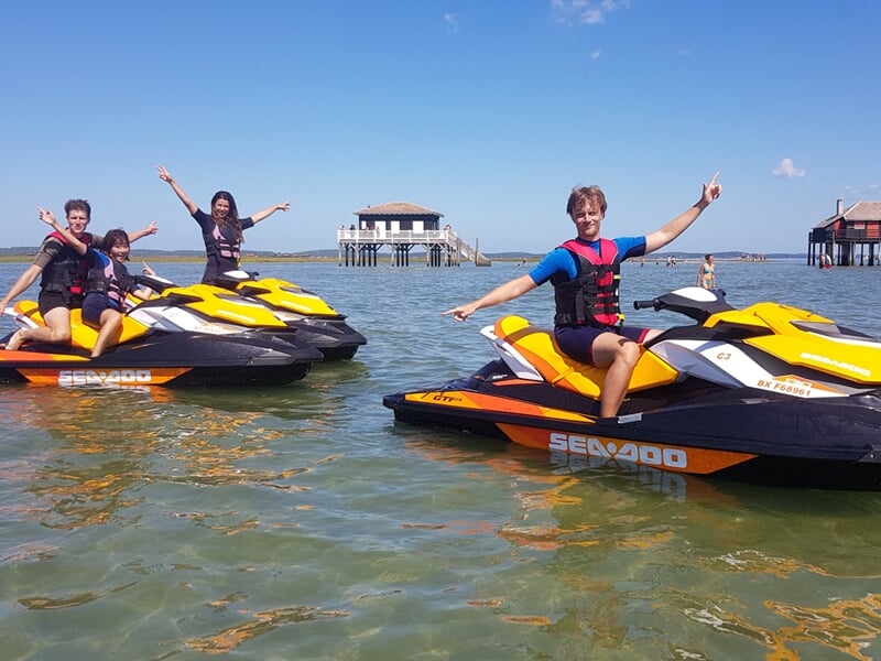 Quatre personnes souriantes sur des jetskis jaunes et noirs dans une eau calme avec une cabane sur pilotis en arrière-plan.