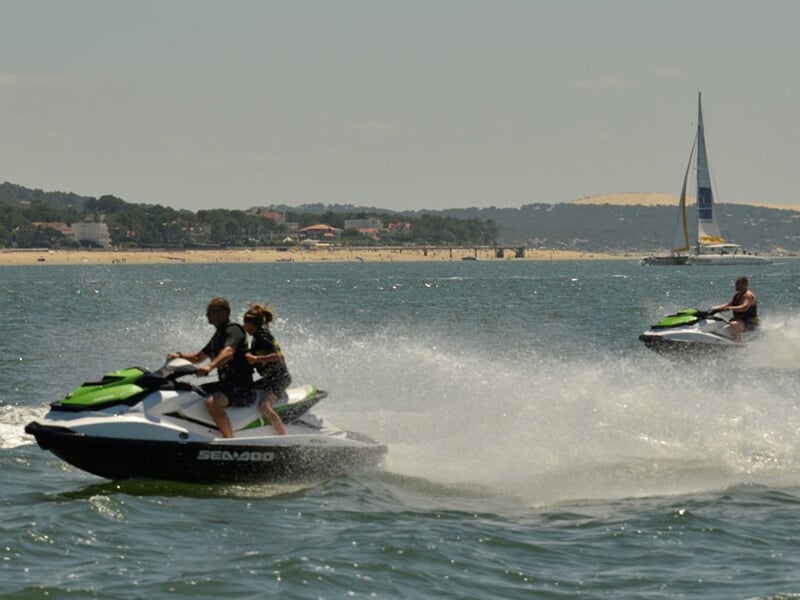 Deux personnes sur un jetski blanc et vert filant sur l'eau avec un autre jetski et un voilier en arrière-plan.