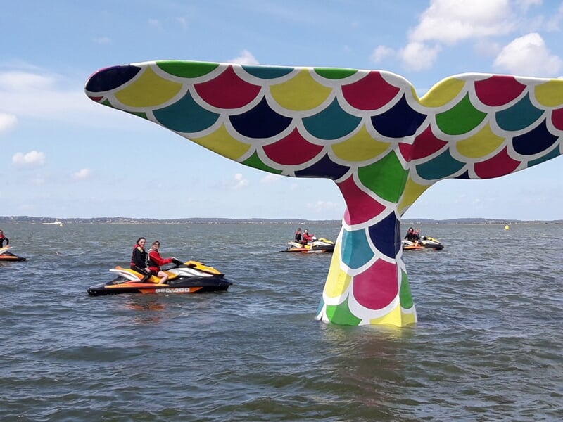 Plusieurs jetskis avec des passagers autour d'une grande sculpture colorée en forme de queue de baleine dans l'eau.